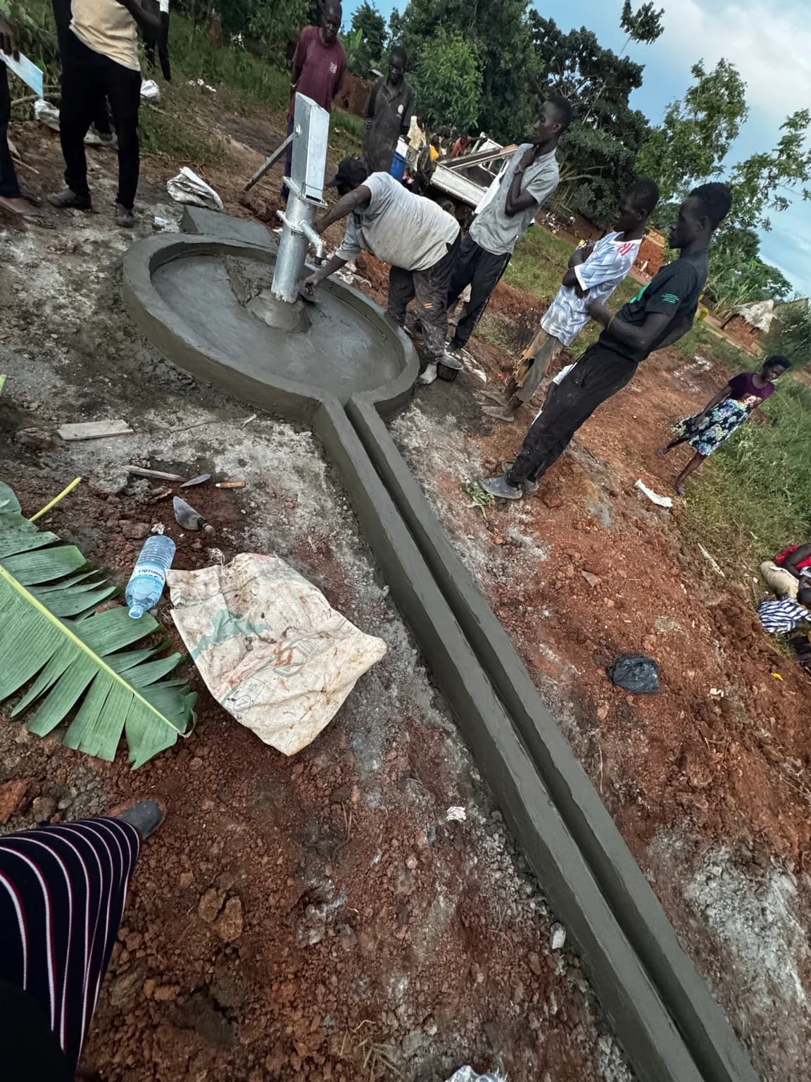A group of men and boys gather around a newly installed hand-pump borehole with a concrete apron and drainage channel in a rural setting.